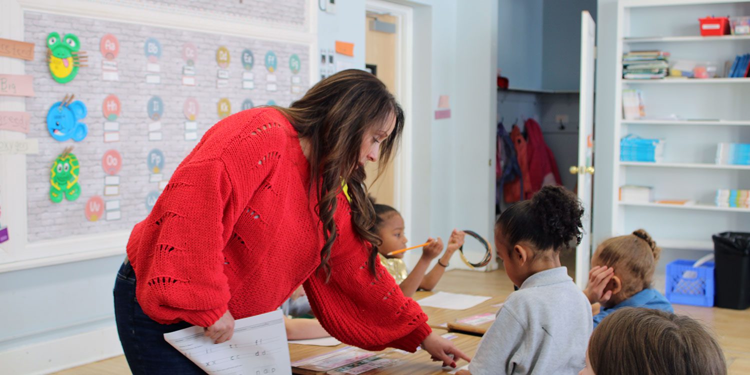 Teacher pointing to a worksheet while helping students at a classroom table.