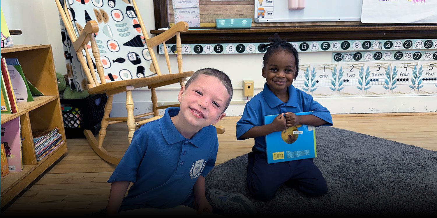 Two smiling students sitting with books on a rug in a classroom.