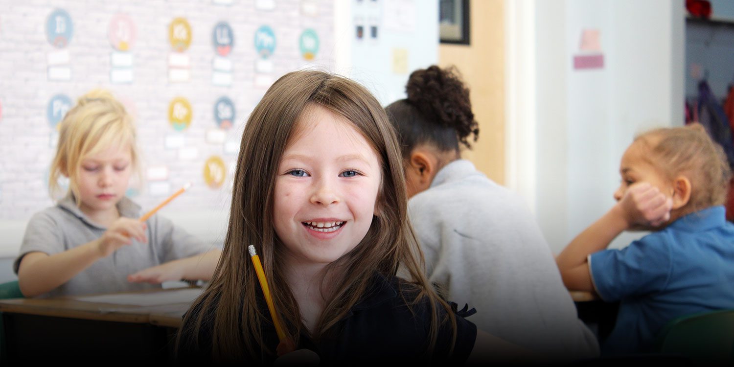 Smiling student holding a pencil at a desk in a Mosaic Classical Academy classroom.